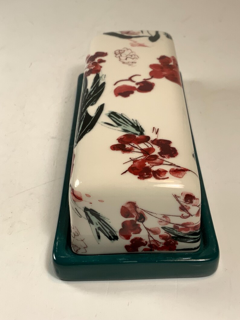 May include: A rectangular butter dish with a white lid featuring a floral design in red and black. The dish sits on a dark teal base. The floral pattern includes red flowers and black leaves.