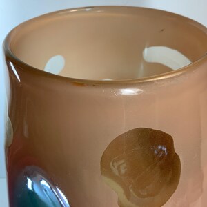 May include: A peach-colored glass vase with a wide opening and a rounded body. The vase has a few white and brown abstract shapes on the exterior. The interior is a lighter peach color. The vase is likely used for decorative purposes.