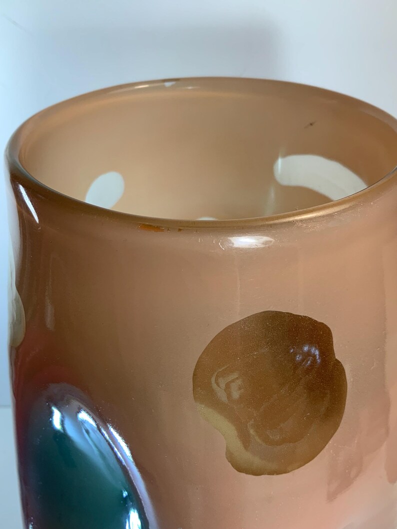 May include: A peach-colored glass vase with a wide opening and a rounded body. The vase has a few white and brown abstract shapes on the exterior. The interior is a lighter peach color. The vase is likely used for decorative purposes.
