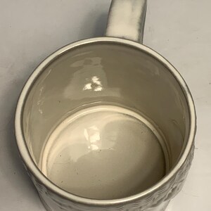 May include: A cream-colored ceramic mug with a rounded body and a sturdy handle. The mug has a textured exterior and a smooth, glossy interior. The handle is rectangular and attached to the side of the mug. The mug is empty.