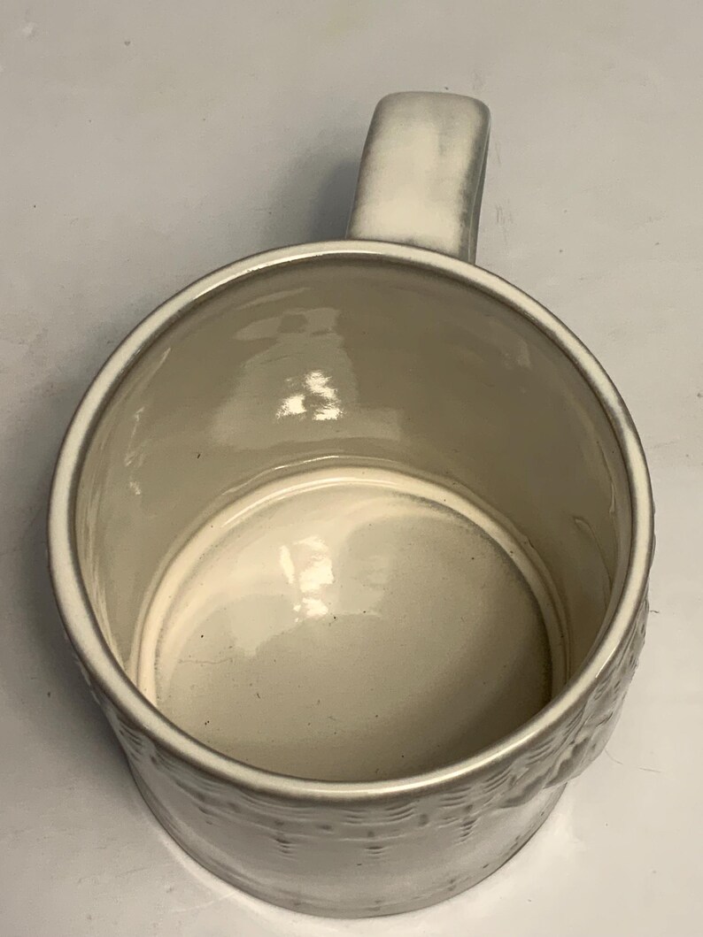 May include: A cream-colored ceramic mug with a rounded body and a sturdy handle. The mug has a textured exterior and a smooth, glossy interior. The handle is rectangular and attached to the side of the mug. The mug is empty.