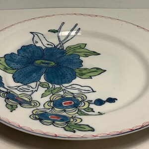 May include: White ceramic plate with a blue floral design. The plate features a large blue flower with green leaves and smaller blue and yellow floral accents. The rim has a red and blue decorative border.