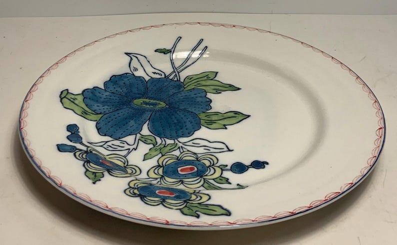 May include: White ceramic plate with a blue floral design. The plate features a large blue flower with green leaves and smaller blue and yellow floral accents. The rim has a red and blue decorative border.