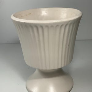May include: A cream-colored, fluted urn-shaped vase with a pedestal base. The vase has a wide opening and vertical grooves along the body. The pedestal base is round and smooth. The vase is made of ceramic or a similar material.