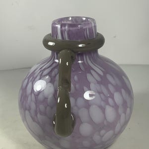 May include: A purple glass vase with white swirls and a brown handle. The vase has a round body and a wide opening.