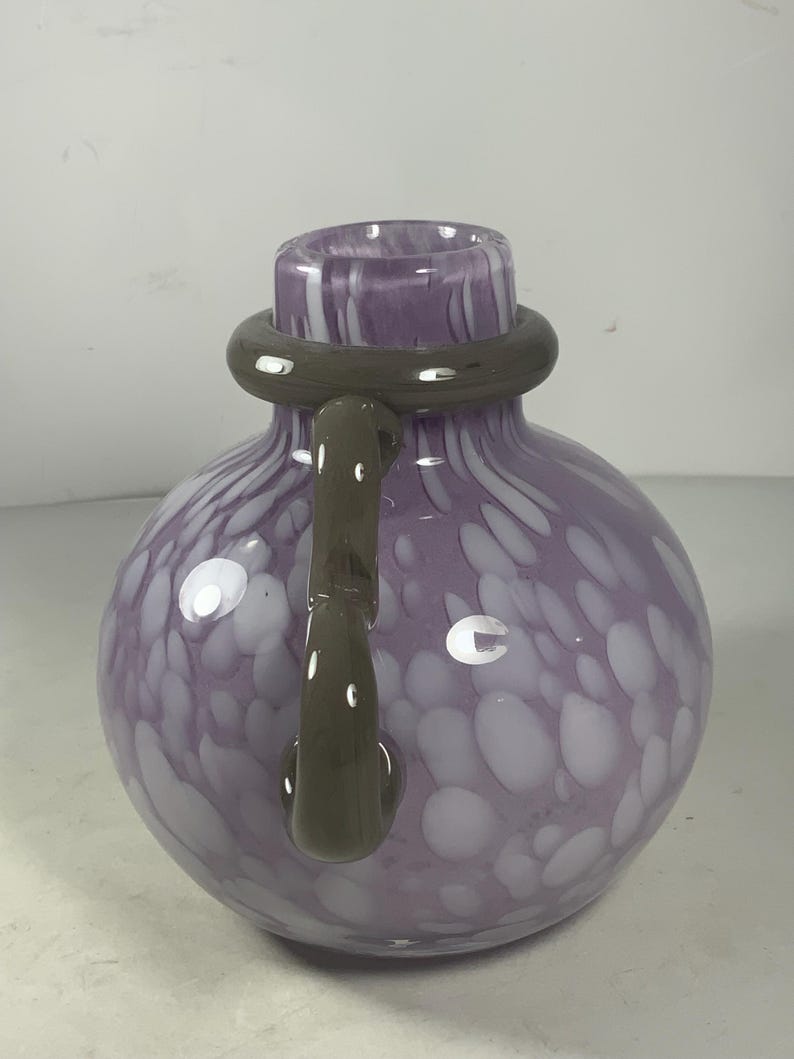 May include: A purple glass vase with white swirls and a brown handle. The vase has a round body and a wide opening.