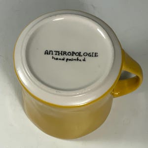 Puede incluir: Una taza de cer&aacute;mica amarilla con interior y asa blancos. La parte inferior de la taza tiene el texto "ANTHROPOLOGIE hand painted" en negro. La taza est&aacute; sobre un fondo blanco.