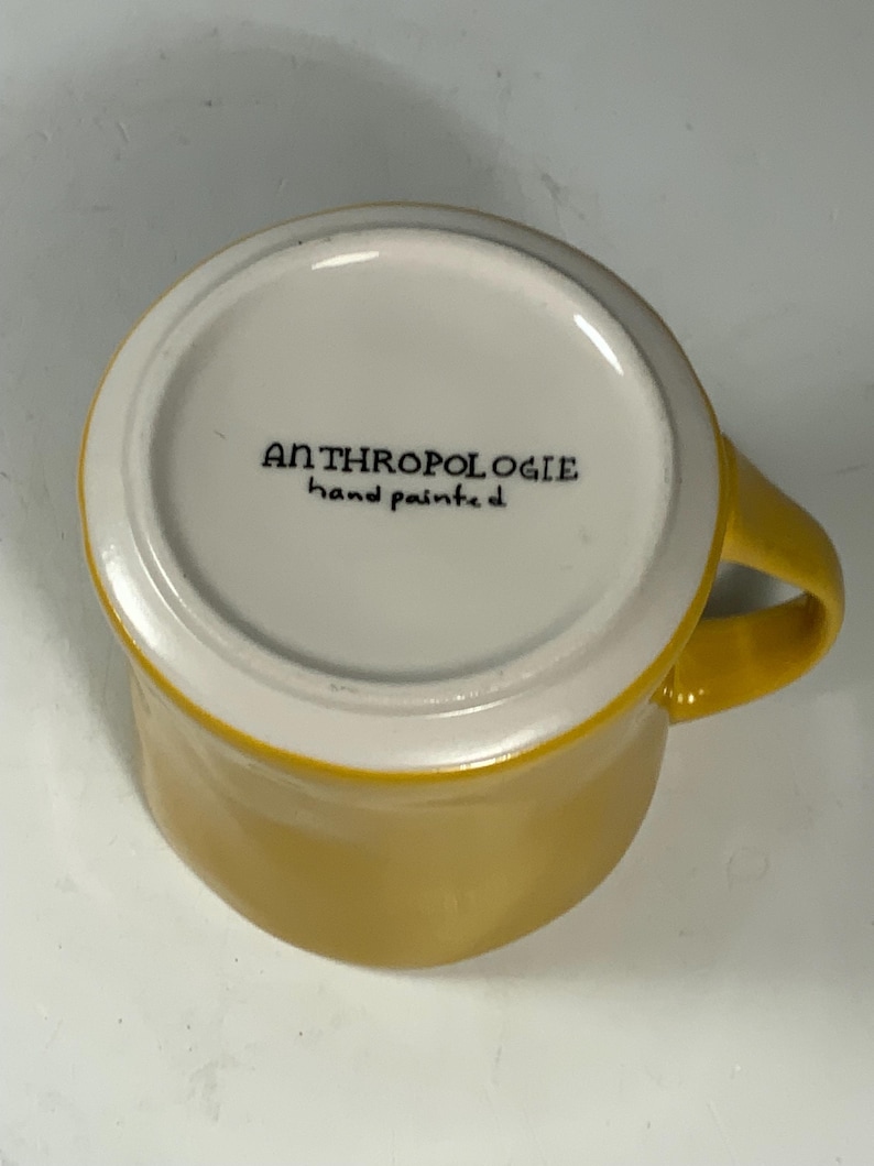 Puede incluir: Una taza de cer&aacute;mica amarilla con interior y asa blancos. La parte inferior de la taza tiene el texto "ANTHROPOLOGIE hand painted" en negro. La taza est&aacute; sobre un fondo blanco.