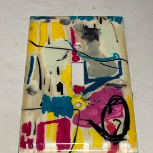 Op de afbeelding: Een rechthoekige wandplaat voor een lichtschakelaar met een glanzende afwerking en een abstract geschilderd ontwerp. Het oppervlak toont levendige spatten en streken van gele, magenta, turquoise, blauwe, grijze en zwarte verf op een crèmekleurige basis.