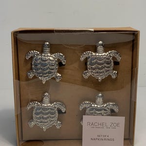 Puede incluir: Un juego de cuatro servilleteros plateados con forma de tortuga, presentados en una caja de cartón con frontal transparente. La caja incluye una etiqueta que dice "RACHEL ZOE" y "SET OF 4 NAPKIN RINGS".
