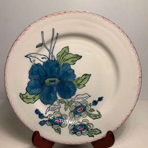 May include: White ceramic plate with a blue floral design. The plate features a large blue flower with green leaves and smaller blue and yellow flowers. The rim has a scalloped red border and a blue line. The plate is displayed on a wooden stand.