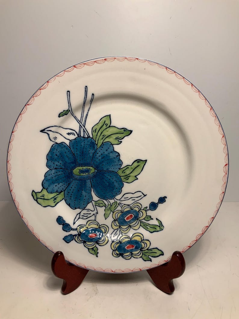 May include: White ceramic plate with a blue floral design. The plate features a large blue flower with green leaves and smaller blue and yellow flowers. The rim has a scalloped red border and a blue line. The plate is displayed on a wooden stand.