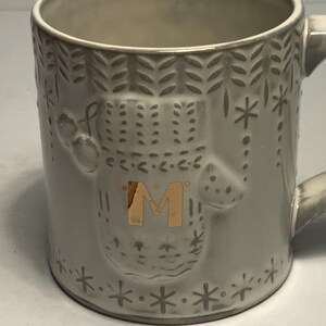 May include: A gray ceramic mug with a decorative design. The mug features an embossed pattern of snowflakes, dots, and a central design with the letter "M" in gold. The mug has a handle and is suitable for hot beverages.