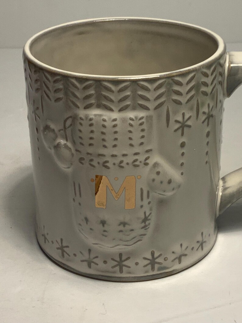 May include: A gray ceramic mug with a decorative design. The mug features an embossed pattern of snowflakes, dots, and a central design with the letter "M" in gold. The mug has a handle and is suitable for hot beverages.