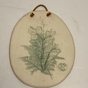 Pode incluir: Pendurador de parede oval em cerâmica branca suja com um design botânico em tons de verde. A obra de arte apresenta ilustrações detalhadas de folhas e fetos. Um cordão de couro castanho está preso para pendurar. Mede cerca de 15 cm de comprimento.