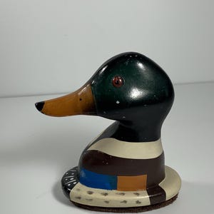 May include: A hand-painted wooden duck figurine. The duck has a black head, a green top, and a brown and orange beak. The body is painted with brown, blue, and cream colors. The duck has a red eye and sits on a brown base.