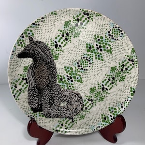May include: Decorative ceramic plate featuring a black and white poodle design. The plate has a white background with a green, black, and blue dotted pattern. The plate is displayed on a dark red wooden stand.