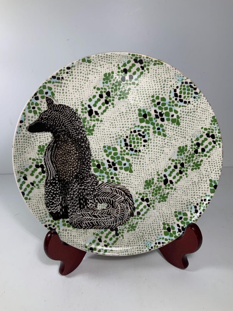 May include: Decorative ceramic plate featuring a black and white poodle design. The plate has a white background with a green, black, and blue dotted pattern. The plate is displayed on a dark red wooden stand.