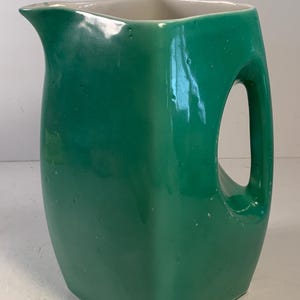 May include: A green ceramic pitcher with a square base and a curved handle. The pitcher has a spout for pouring and a white interior. The exterior is a glossy, solid green color.