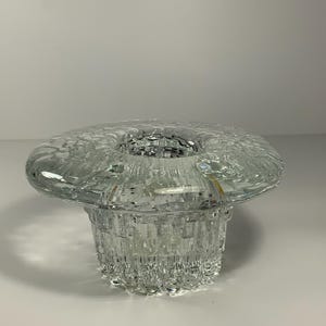 Puede incluir: Un portavelas de cristal transparente con una parte superior ancha y texturizada y una base cilíndrica. La base tiene un patrón vertical acanalado y la parte superior tiene un diseño circular texturizado. El portavelas está diseñado para una sola vela.