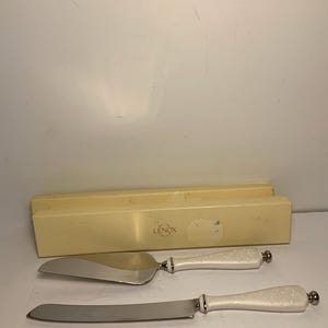 May include: A cake server and knife set with white handles and silver blades. The handles have a dotted pattern and silver accents. The set is from Lenox and comes with a cream-colored box.