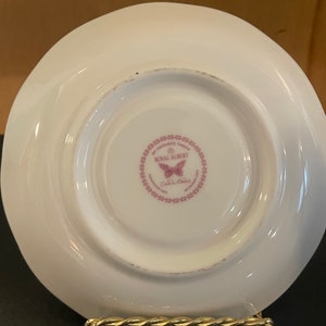 May include: White porcelain saucer with a scalloped edge, displaying a pink Royal Albert logo with a butterfly and the words "My Favourite Things". The saucer is on a gold-colored metal stand.