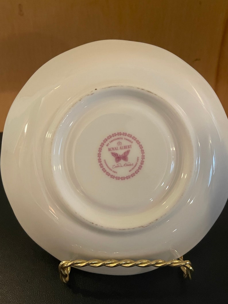 May include: White porcelain saucer with a scalloped edge, displaying a pink Royal Albert logo with a butterfly and the words "My Favourite Things". The saucer is on a gold-colored metal stand.