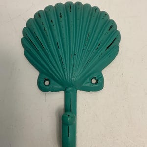 May include: A teal seashell-shaped wall hook with a distressed finish. The hook has a single prong and two holes for mounting. The metal hook is designed to resemble a scallop shell.