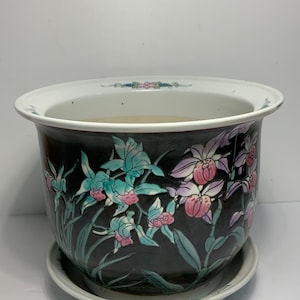 Könnte beinhalten: Schwarzer Keramik-Blumentopf mit weißem Rand und passender Untertasse. Der Topf ist mit einem Blumendesign in Rosa, Türkis und Grün verziert. Der Topf ist rund und hat eine weite Öffnung.