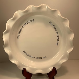 May include: A white ceramic pie dish with a ruffled edge, resting on a dark brown stand. The dish has the phrase "Pie fixes everything" printed in black around the rim. The dish is designed for baking and serving pies.