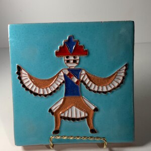 Peut inclure: Carreau de céramique turquoise représentant une figure stylisée aux ailes déployées. La figure porte une coiffe rouge et bleue, un haut bleu et une jupe beige. Les ailes sont beiges avec des accents blancs et noirs. La tuile est exposée sur un support en fil d'or.