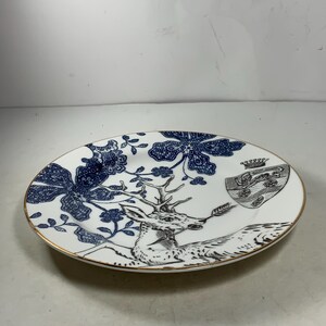 May include: A white ceramic plate with a blue and gold design. The plate features a deer with antlers, a floral pattern, and a crest with a crown.