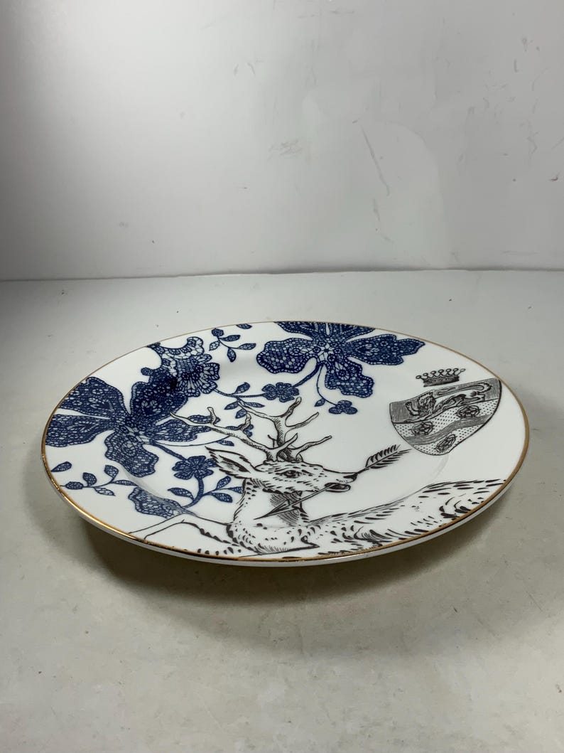 May include: A white ceramic plate with a blue and gold design. The plate features a deer with antlers, a floral pattern, and a crest with a crown.