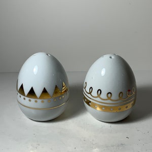 Op de afbeelding: Een set van twee witte, eivormige zout- en peperstrooiers van keramiek met gouden accenten. De ene strooier heeft een geometrisch ontwerp en de andere heeft een golvend lijnenontwerp.