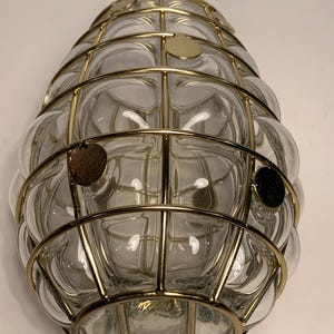 Op de afbeelding: Een heldere glazen lamp met een bubbelachtig ontwerp, omhuld door een goudkleurige metalen kooi. De lamp heeft een afgeronde vorm en is aan een muur bevestigd. De metalen kooi heeft een rasterpatroon en de glazen bellen zijn zichtbaar.