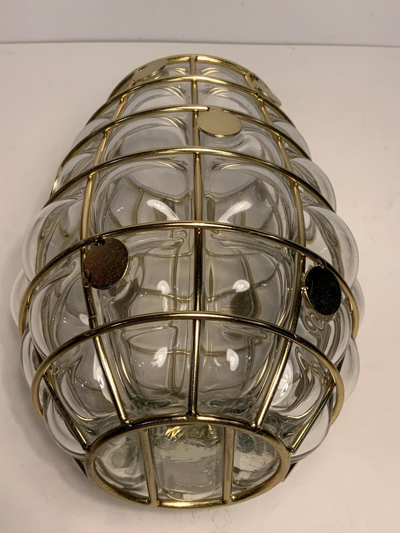 Op de afbeelding: Een heldere glazen lamp met een bubbelachtig ontwerp, omhuld door een goudkleurige metalen kooi. De lamp heeft een afgeronde vorm en is aan een muur bevestigd. De metalen kooi heeft een rasterpatroon en de glazen bellen zijn zichtbaar.