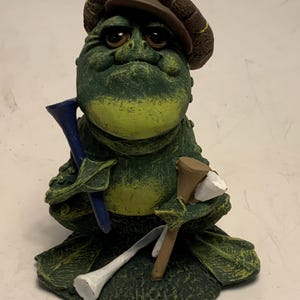 May include: A whimsical ceramic frog figurine wearing a brown cap, holding golf tees. The frog is primarily green with yellow accents, and the tees are blue, brown, and white. The figurine has a detailed, textured finish.