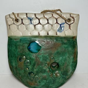 Op de afbeelding: Een keramische wandzak met een platte achterkant en een afgeronde onderkant. Het bovenste gedeelte heeft een wit honingraatpatroon met bruine omlijningen, terwijl het onderste gedeelte een gevlekte groene glazuur heeft met een gat en blauwe accenten.