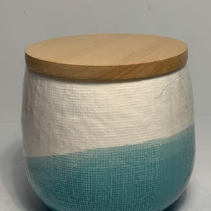 May include: A ceramic jar with a wooden lid. The jar is white on top and transitions to a teal blue colour on the bottom. The jar has a textured surface.