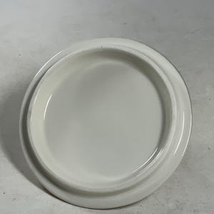 Może przedstawiać: Biała ceramiczna pokrywka o gładkiej, zaokrąglonej powierzchni. Pokrywka jest zaprojektowana do dopasowania do pojemnika lub słoika.
