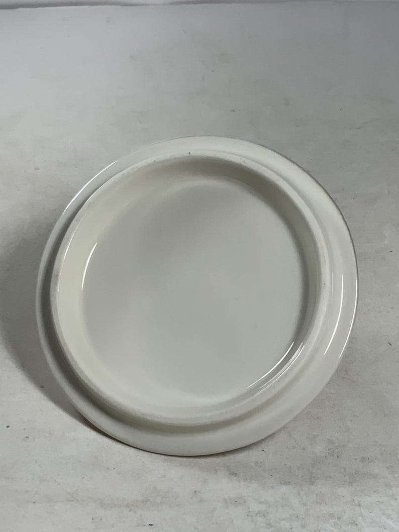 Może przedstawiać: Biała ceramiczna pokrywka o gładkiej, zaokrąglonej powierzchni. Pokrywka jest zaprojektowana do dopasowania do pojemnika lub słoika.