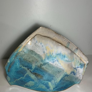 May include: A ceramic napkin holder with a triangular, mountain-like silhouette. The surface features a reactive glaze with blended shades of deep ocean blue, turquoise, soft green, and creamy white, creating a fluid, marbled appearance.