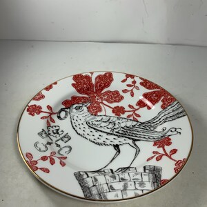 May include: A white porcelain plate with a red and black floral design. A black and white bird with a ring in its beak is perched on a brick structure. The plate has a gold rim.