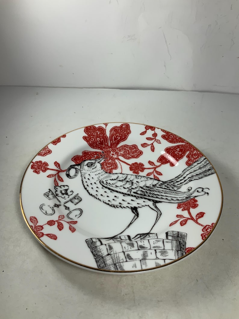 May include: A white porcelain plate with a red and black floral design. A black and white bird with a ring in its beak is perched on a brick structure. The plate has a gold rim.