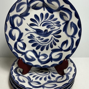 May include: A set of four round ceramic plates featuring a hand-painted dark blue floral and leaf pattern on a white background. The center of each plate displays a stylized bird motif surrounded by a circular arrangement of brushstroke leaves.