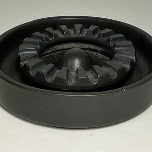 May include: A circular matte black ashtray featuring a raised central dome surrounded by a gear-shaped ring with multiple notches. The design includes a deep outer basin and a smooth, uniform finish across the entire structure of the object.