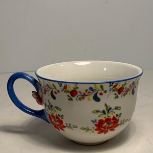 May include: A white ceramic teacup with a blue handle and rim. The cup is decorated with a floral pattern in red, green, and yellow. The handle has a small red flower attached.