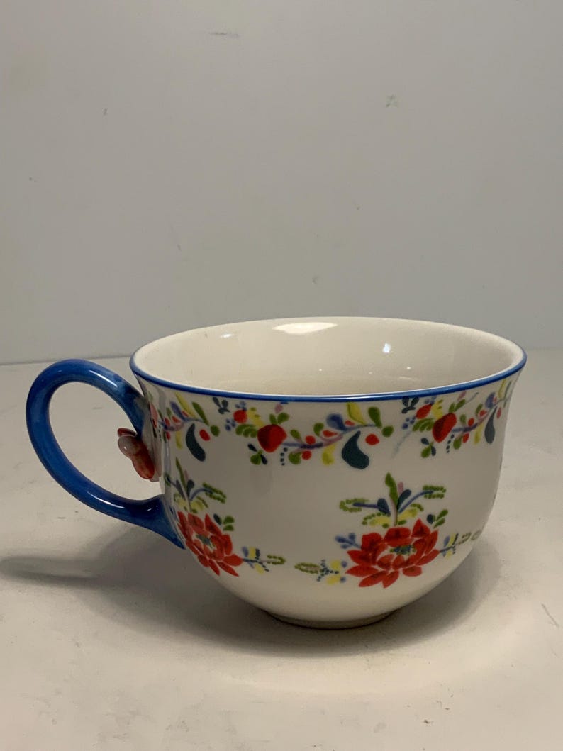 May include: A white ceramic teacup with a blue handle and rim. The cup is decorated with a floral pattern in red, green, and yellow. The handle has a small red flower attached.