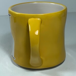 Puede incluir: Una taza de cer&aacute;mica amarilla con un asa curva. El interior de la taza es blanco. La taza tiene un acabado brillante y parece estar vac&iacute;a. La taza est&aacute; sobre una superficie blanca.
