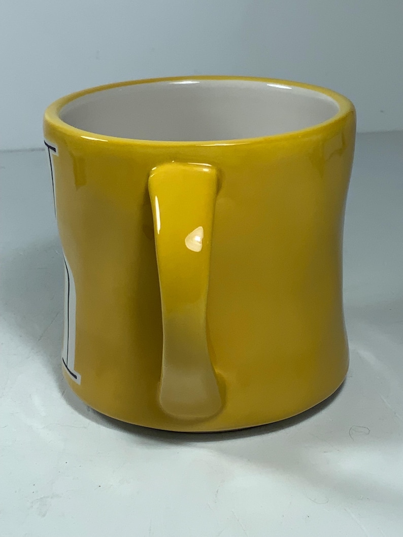 Puede incluir: Una taza de cer&aacute;mica amarilla con un asa curva. El interior de la taza es blanco. La taza tiene un acabado brillante y parece estar vac&iacute;a. La taza est&aacute; sobre una superficie blanca.